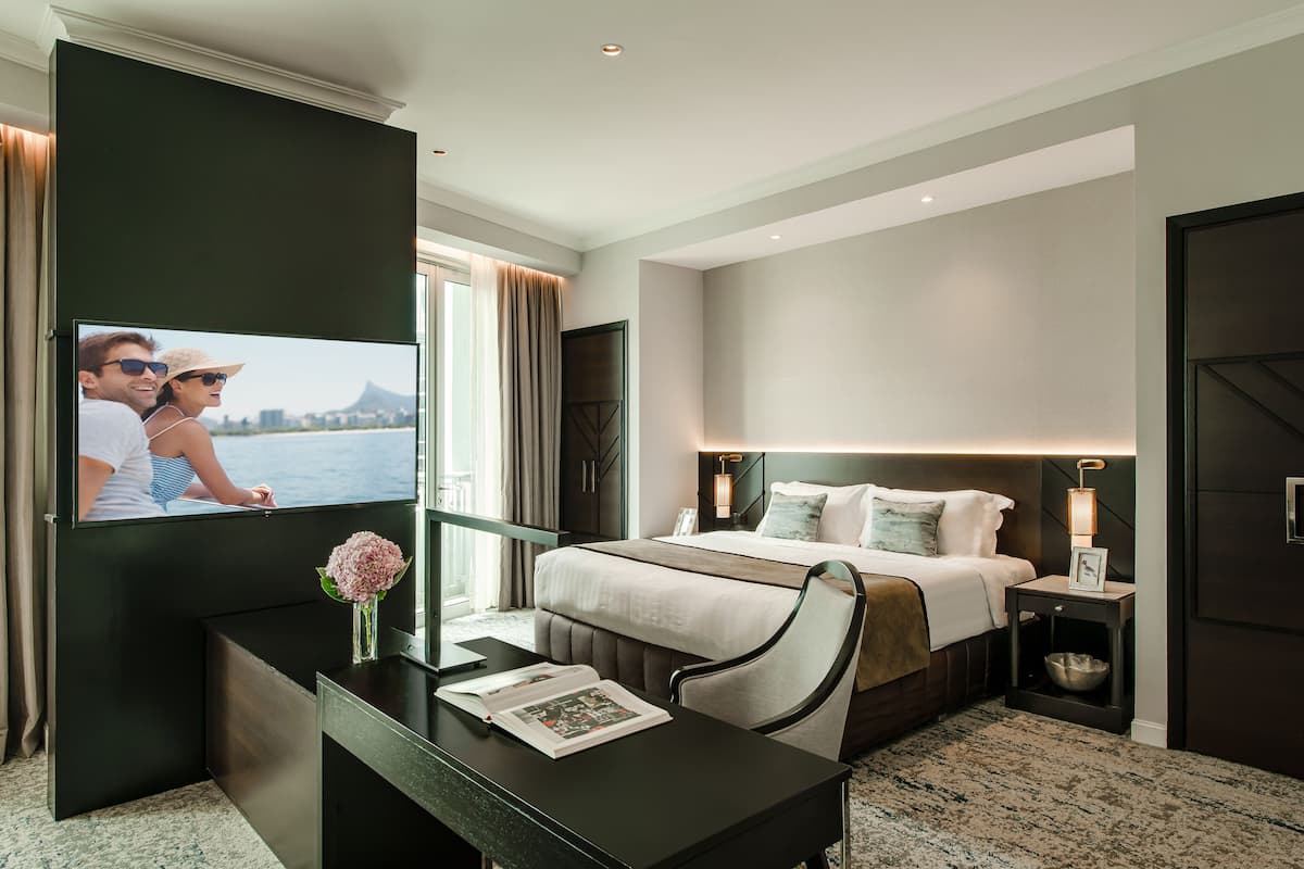 Une chambre d’hôtel moderne avec un grand lit, un bureau avec un livre, une télévision et une vue sur une étendue d’eau.