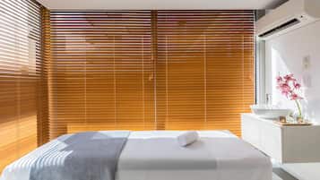 A modern hotel room with a large bed, wooden blinds, a white bedside table with a vase of flowers, and a view of the outdoors.