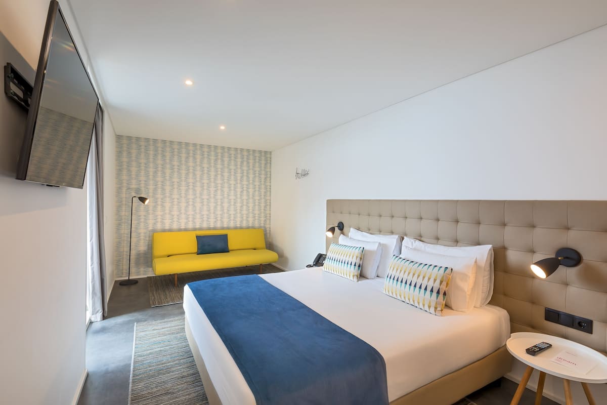 A hotel room with a bed, a yellow sofa, a TV, and a bedside table.