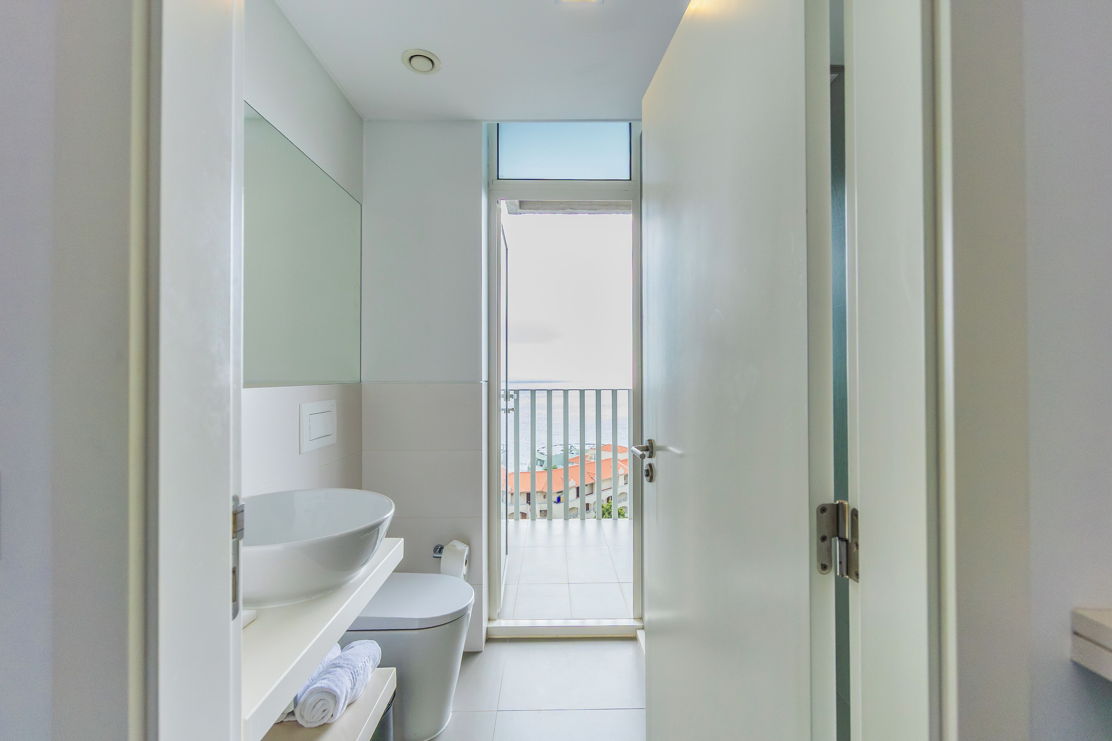 A modern bathroom with a white sink, toilet, and a glass door leading to a balcony.