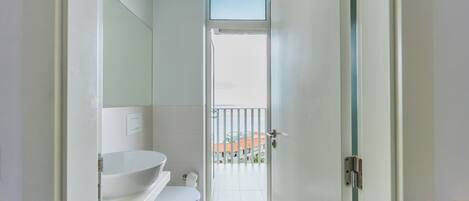 Une salle de bain moderne avec un lavabo blanc, des toilettes et une porte en verre donnant sur un balcon.