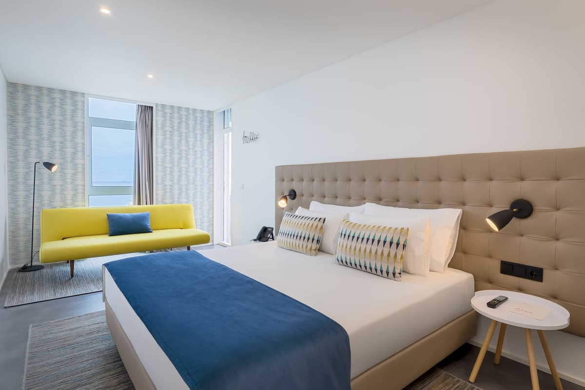 A modern hotel room with a large bed, a yellow sofa, and a small bedside table.