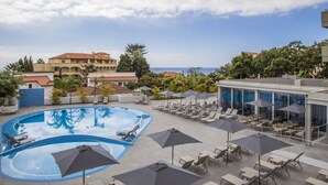 Zona de piscina con sillones de descanso y sombrillas, vista despejada al mar y un edificio con tejado rojo al fondo.