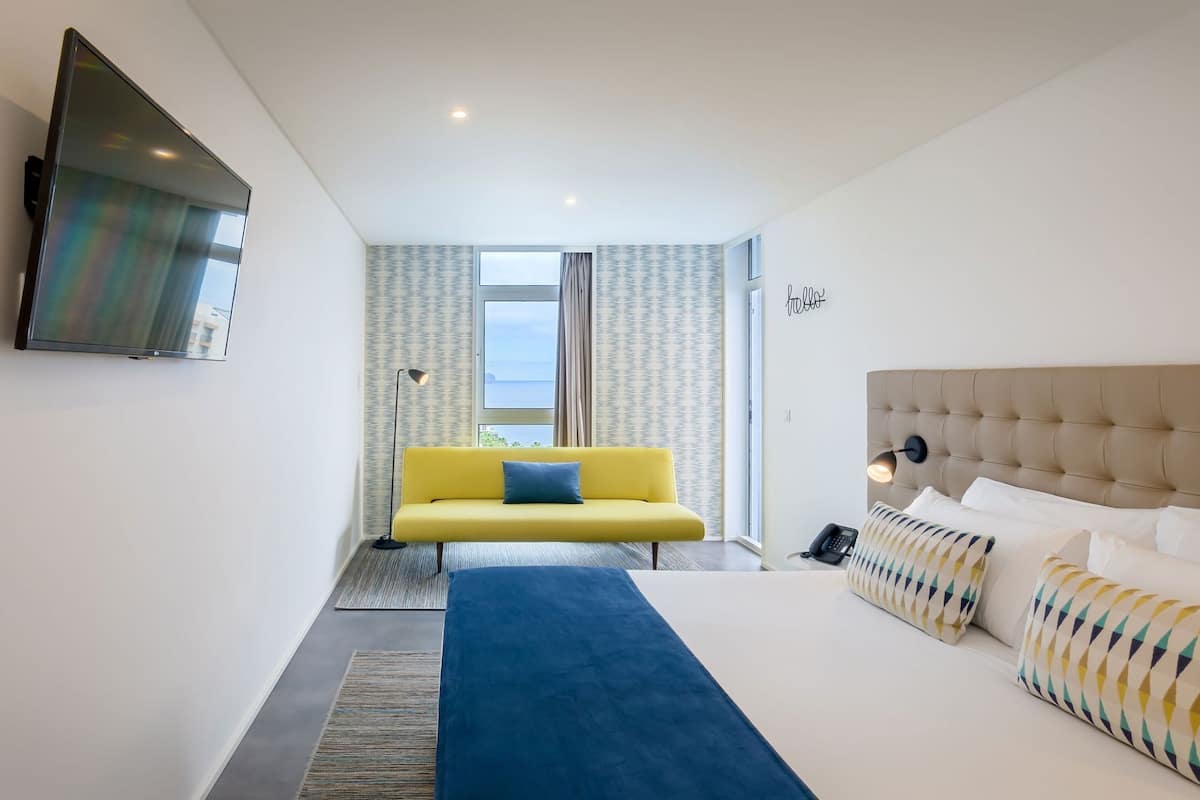 A hotel room with a bed, a yellow sofa, a TV, and a view of the sea.
