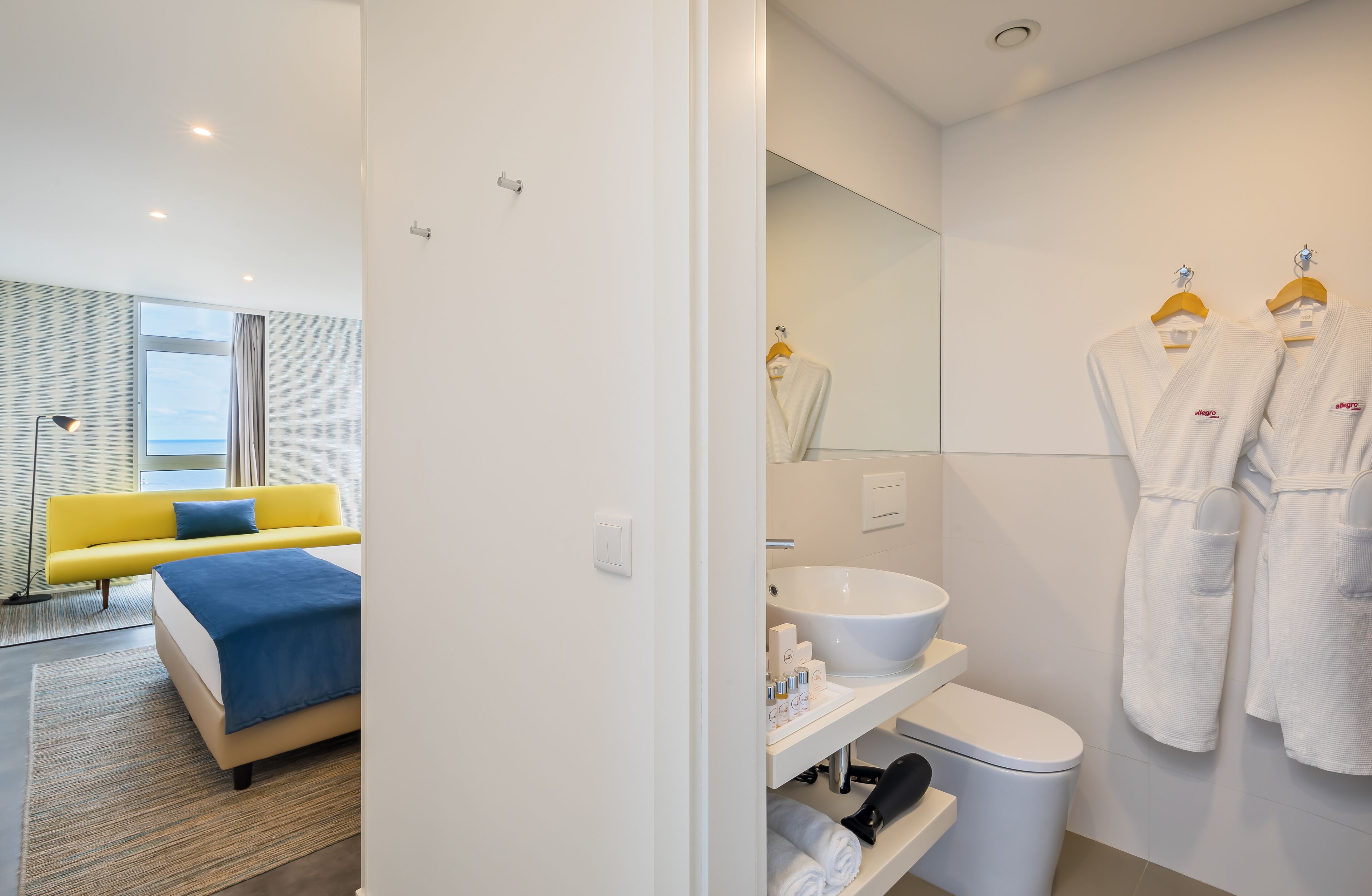 A hotel room with a bed, a yellow sofa, a large window, a bathroom with a sink, toilet, and two white robes hanging on the wall.