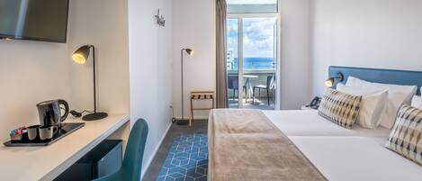 A hotel room with a bed, desk, chair, and a view of the sea through a large window.