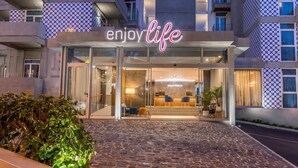 Entrada de hotel con un letrero de neón que dice "enjoy life", una recepción y un camino empedrado.