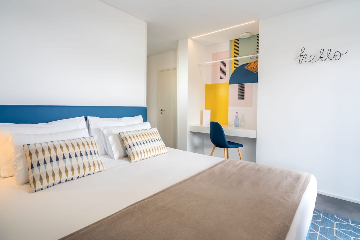 A modern hotel room with a neatly made bed, a desk with a chair, and a 'hello' sign on the wall.