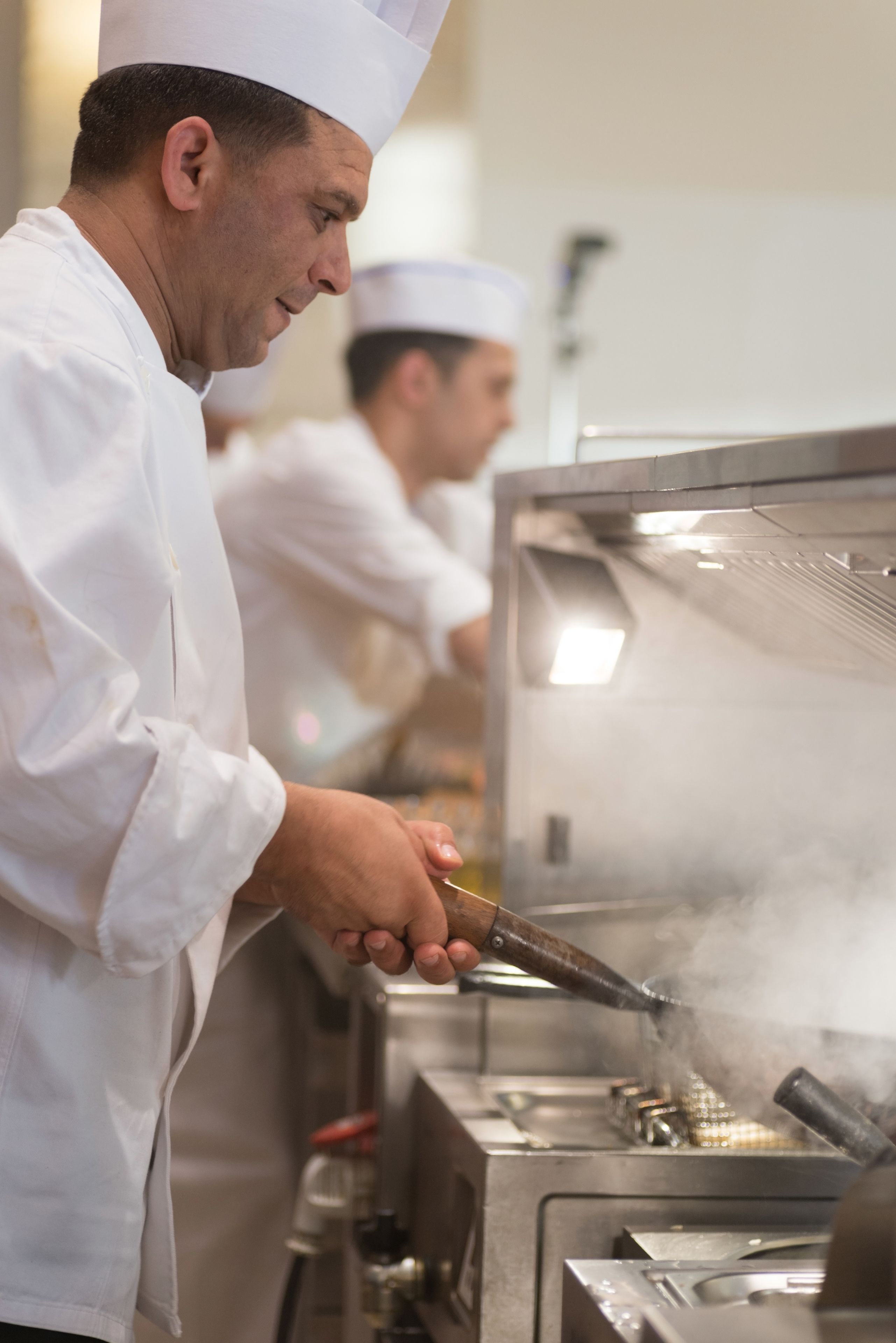 A chef in a white uniform and hat cooking with a pan, steam rising from the pan, in a professional kitchen setting.