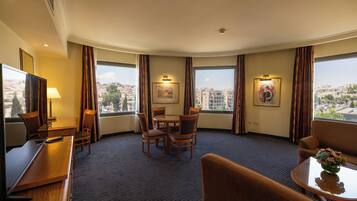 A hotel room with a curved layout, a dining area with a table and chairs, a sofa, a TV, and large windows with curtains.
