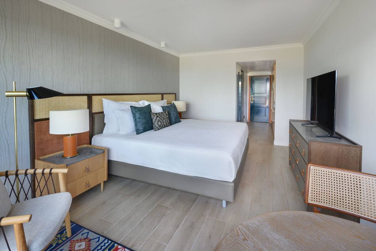 Ein modernes Hotelzimmer mit einem großen Bett, einem Fernseher, einem Nachttisch aus Holz, einem Sessel und einem Schminktisch.