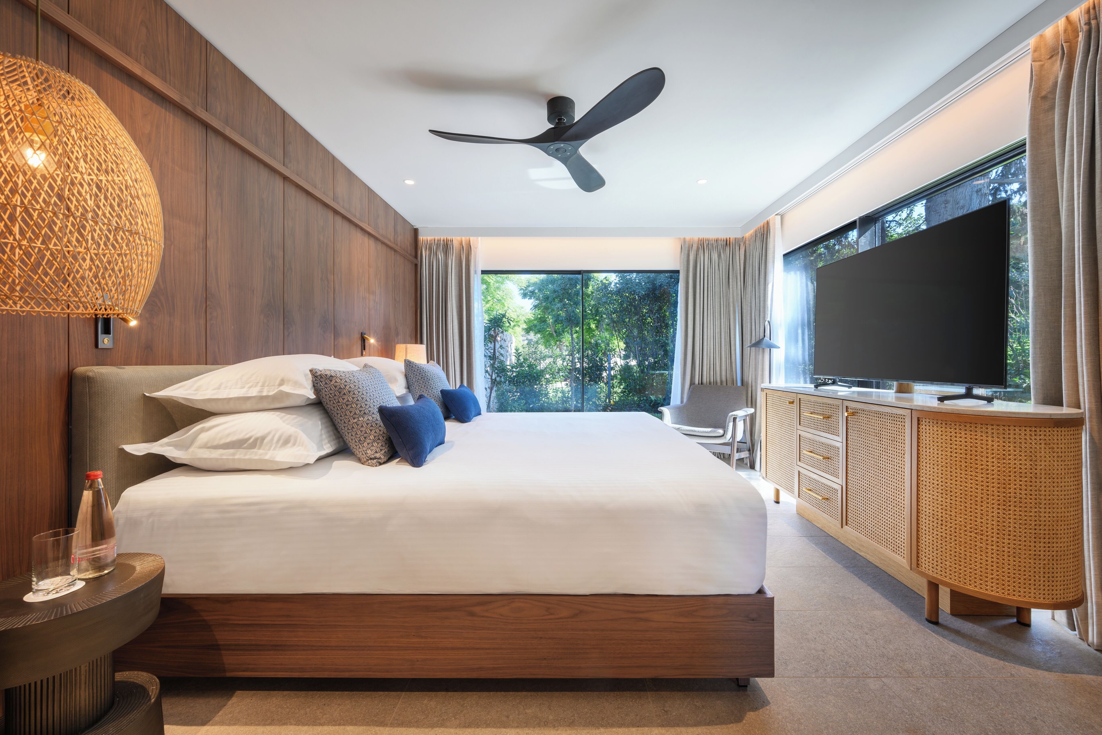 Ein modernes Schlafzimmer mit einem großen Bett, einem Deckenventilator, einem Fernseher und Blick auf Grünflächen.
