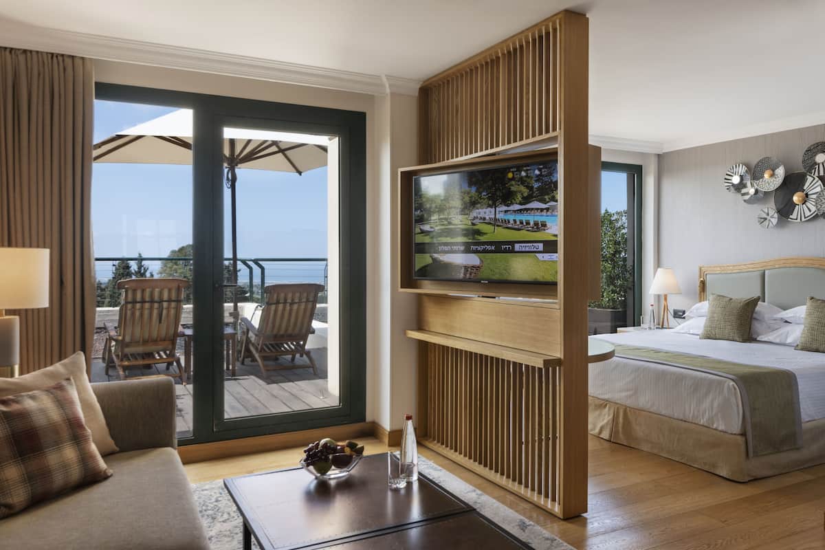 Ein Hotelzimmer mit Bett, Fernseher an einer Holzverkleidung, Balkon mit Sitzmöbeln und Meerblick.