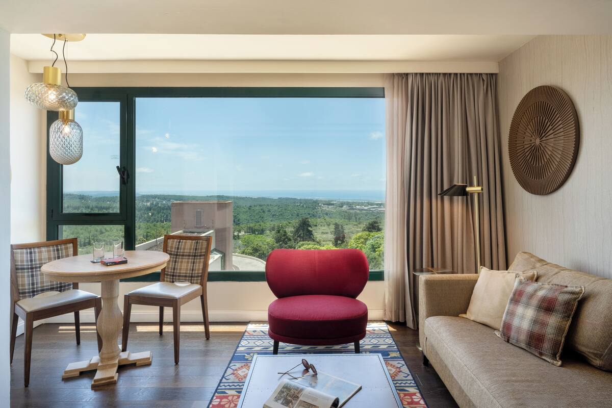Ein Zimmer mit einem großen Fenster, einem roten Sessel, einer Couch, einem Tisch mit Gläsern und einem gemusterten Teppich.