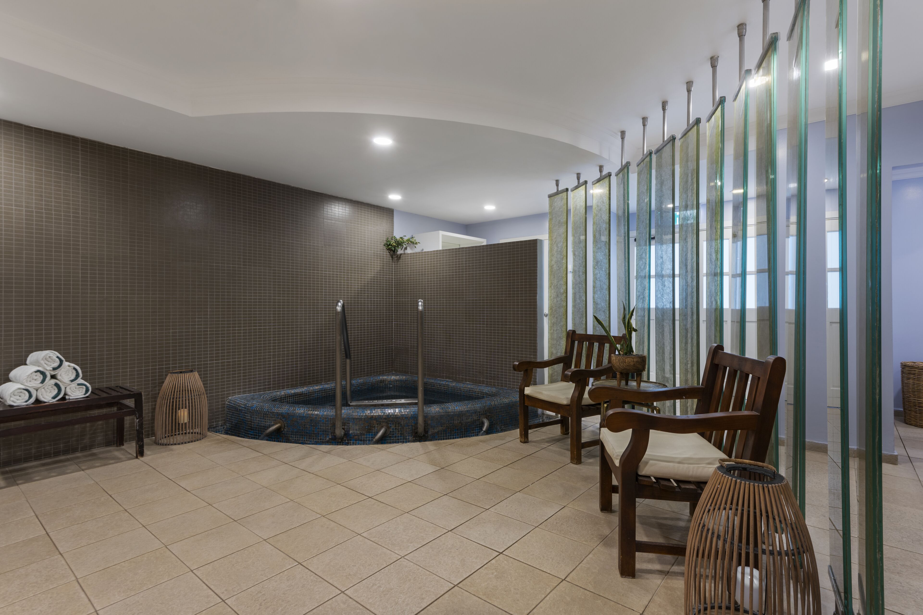 Un espace spa comprenant un jacuzzi, des chaises en bois, un banc avec des serviettes et une cloison en verre.