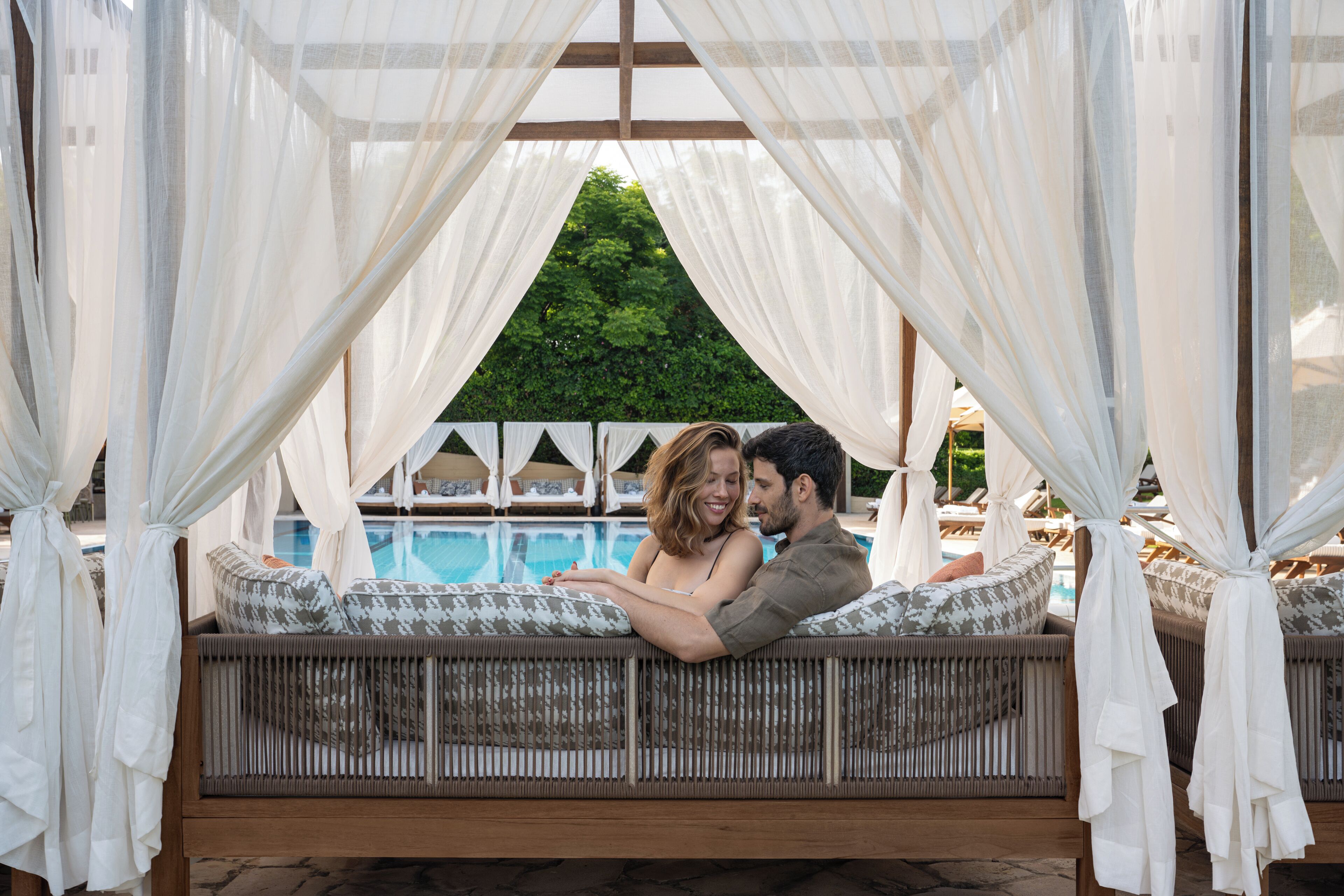 Una pareja sentada en un sofá de mimbre con dosel, con vistas a una piscina y follaje.