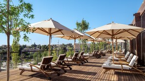 A wooden deck with lounge chairs and umbrellas, overlooking a cityscape.