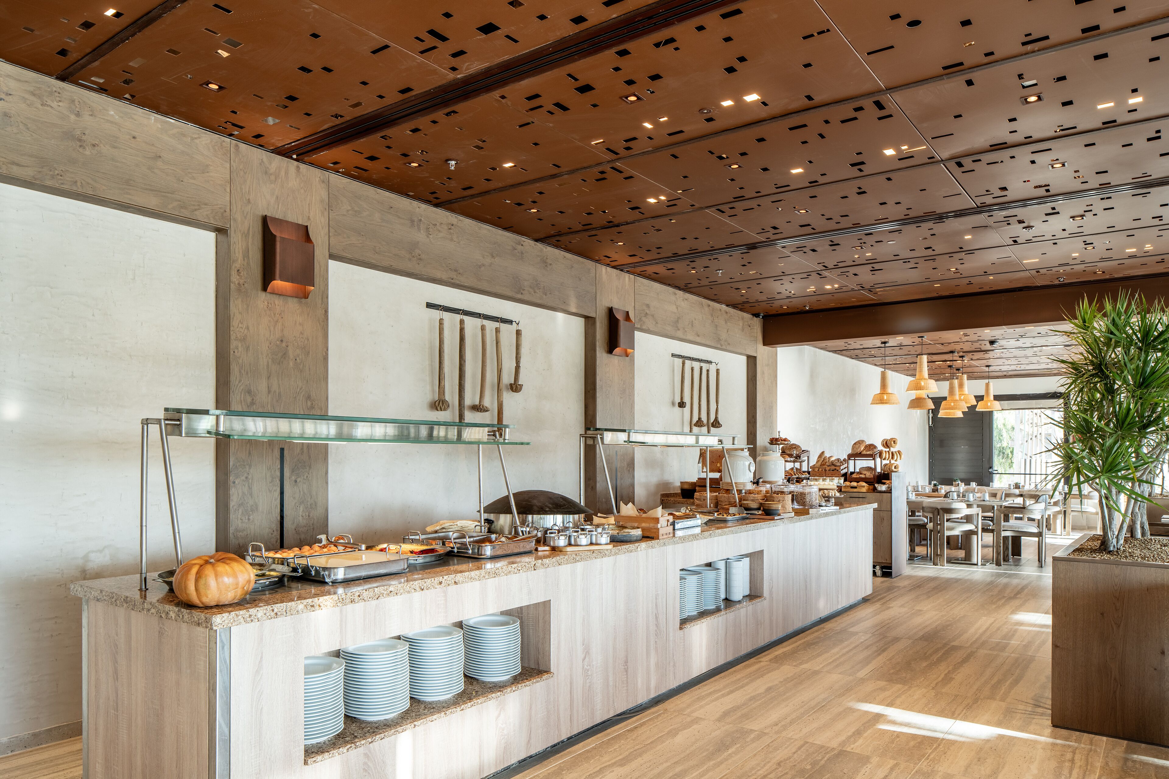 Una barra de desayuno moderna con diversos alimentos, techo de madera con vigas a la vista y un área de estar con mesas y sillas.