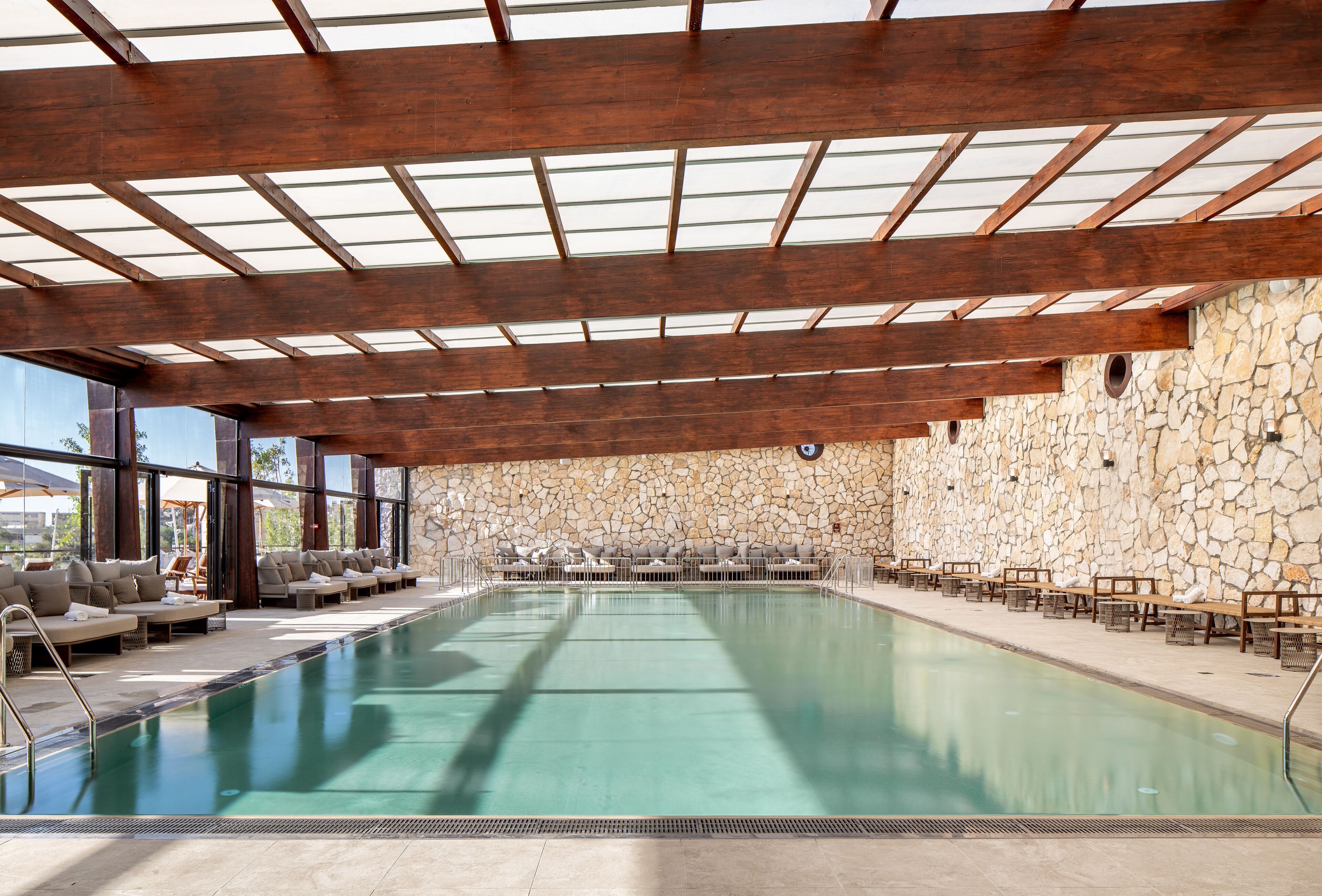 Una amplia piscina interior con pared de piedra y vigas de madera en el techo.