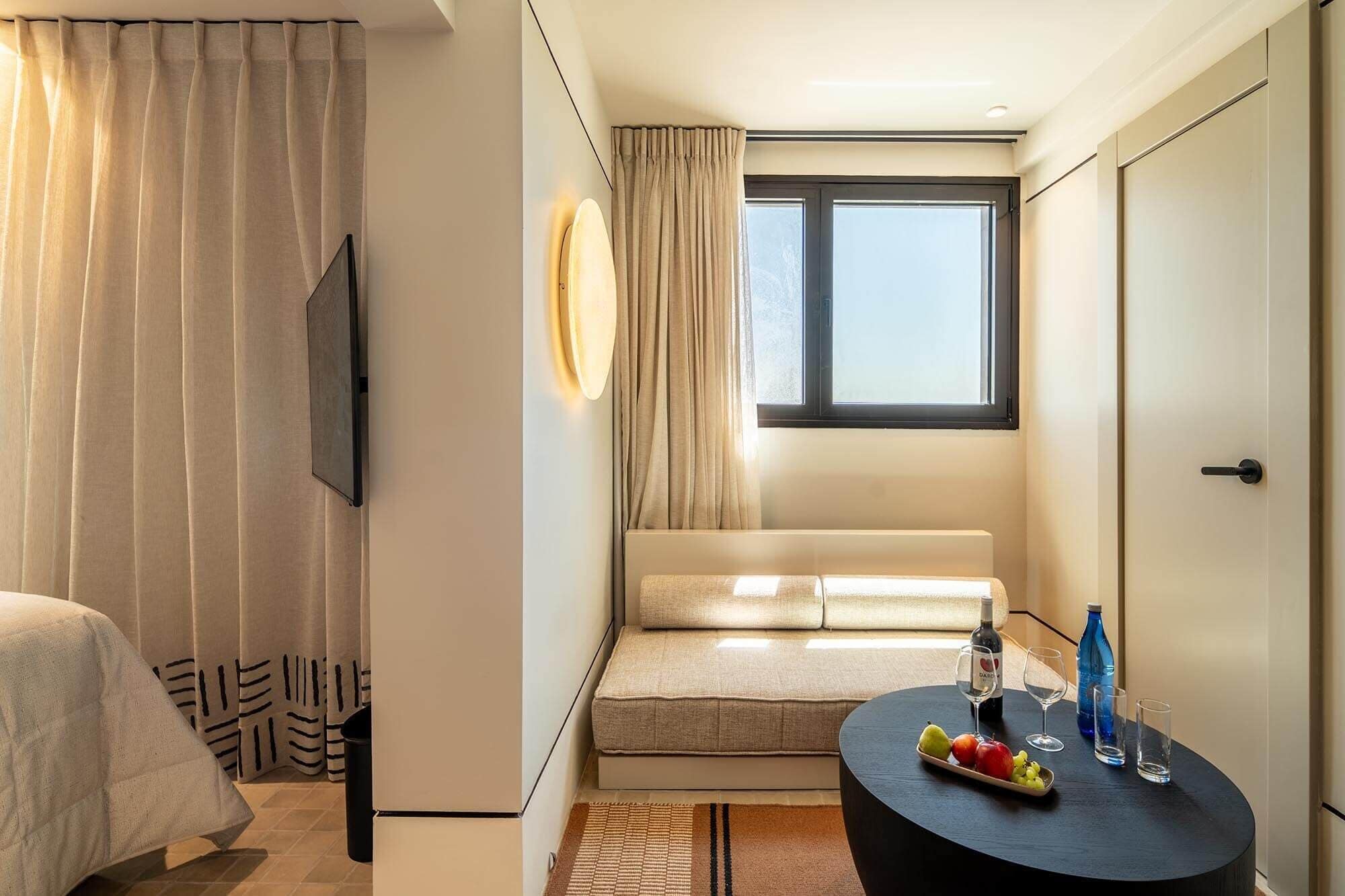 Ein Hotelzimmer mit Bett, einer Couch, einem Esstisch mit Früchten und Getränken, einem Fernseher und einem Fenster mit Vorhängen.
