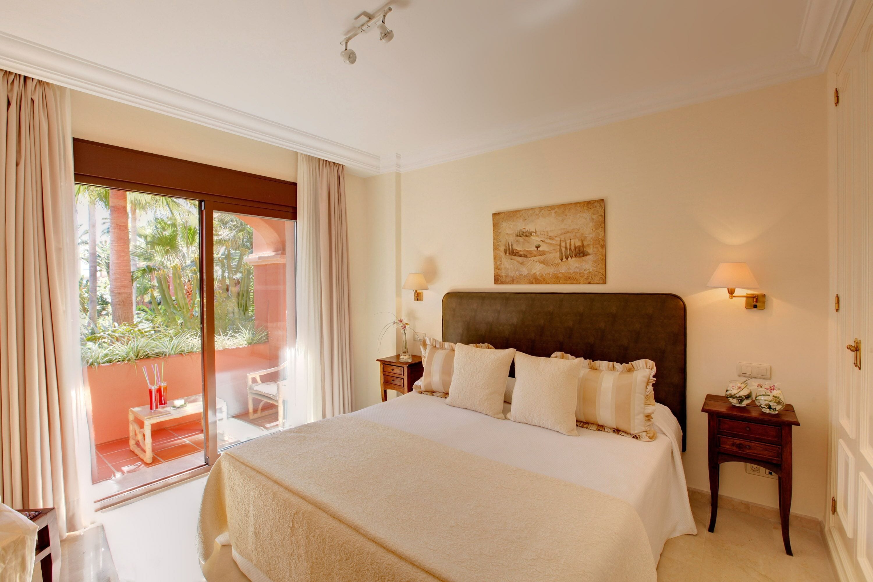 A hotel room with a large bed, a nightstand, a lamp, a painting on the wall, and a view of the outdoors through a sliding door.
