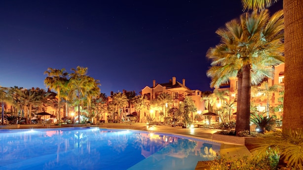 Vista nocturna de un resort con piscina, palmeras y edificios iluminados.