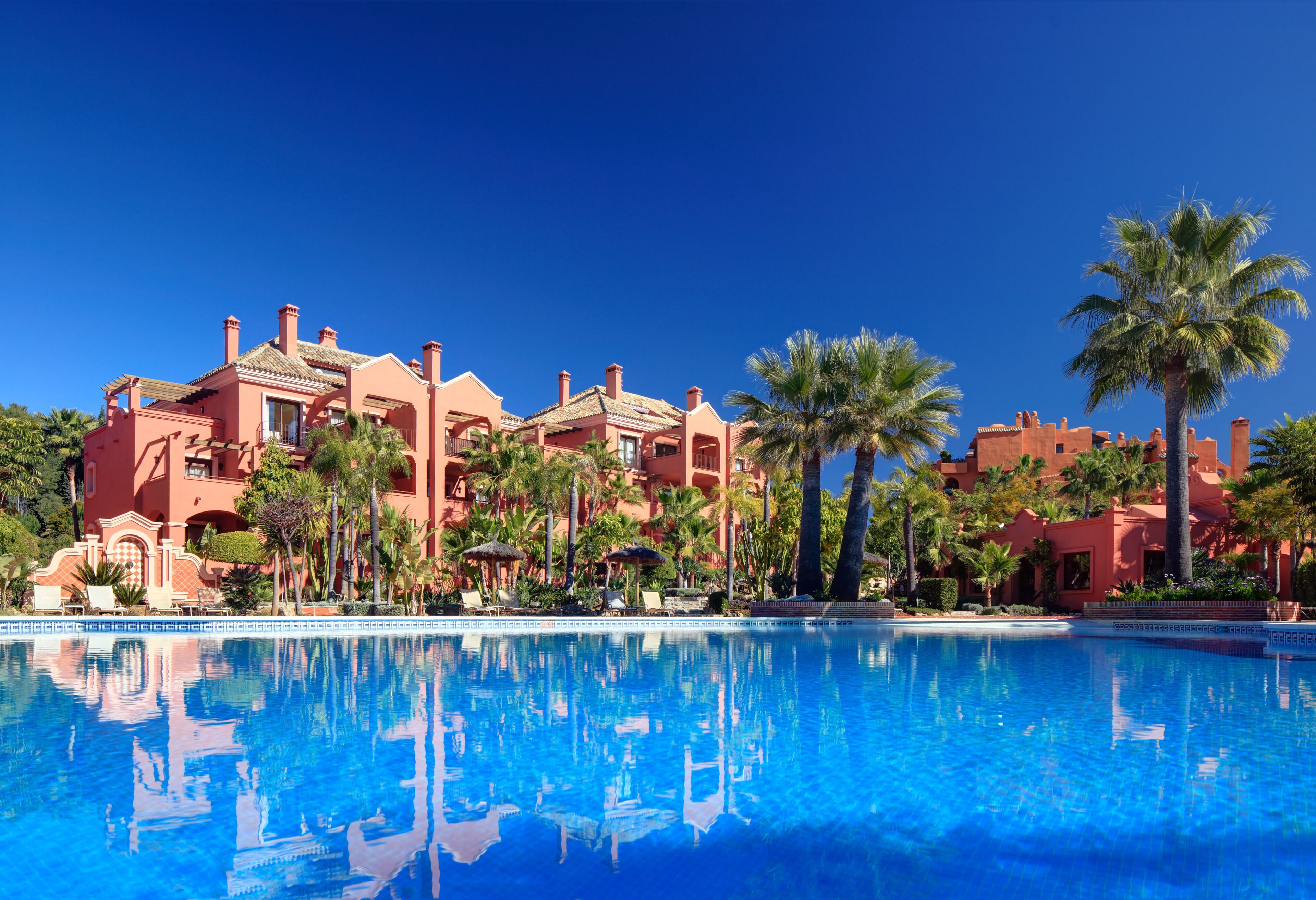 A hotel with a large pool, palm trees, and a clear blue sky.