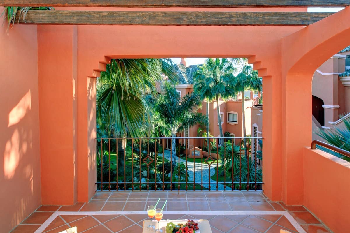 A covered patio with wicker chairs, a table with fruit and drinks, and a view of a swimming pool and palm trees.