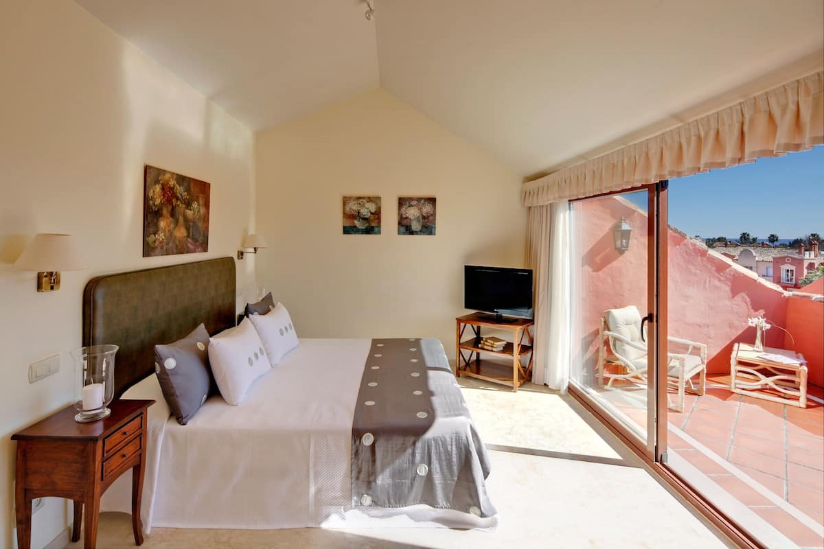 A bedroom with a bed, bedside table, TV, and a balcony with chairs and a table.