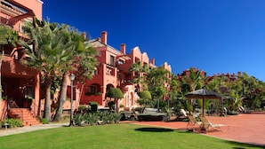 Un hotel con fachada roja, palmeras y una zona de piscina con sillones de descanso.