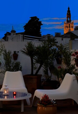 An outdoor seating area with white chairs and a round table, surrounded by potted plants and a view of a historic tower at dusk. - Hotel Un Patio en Santa Cruz (Seville)