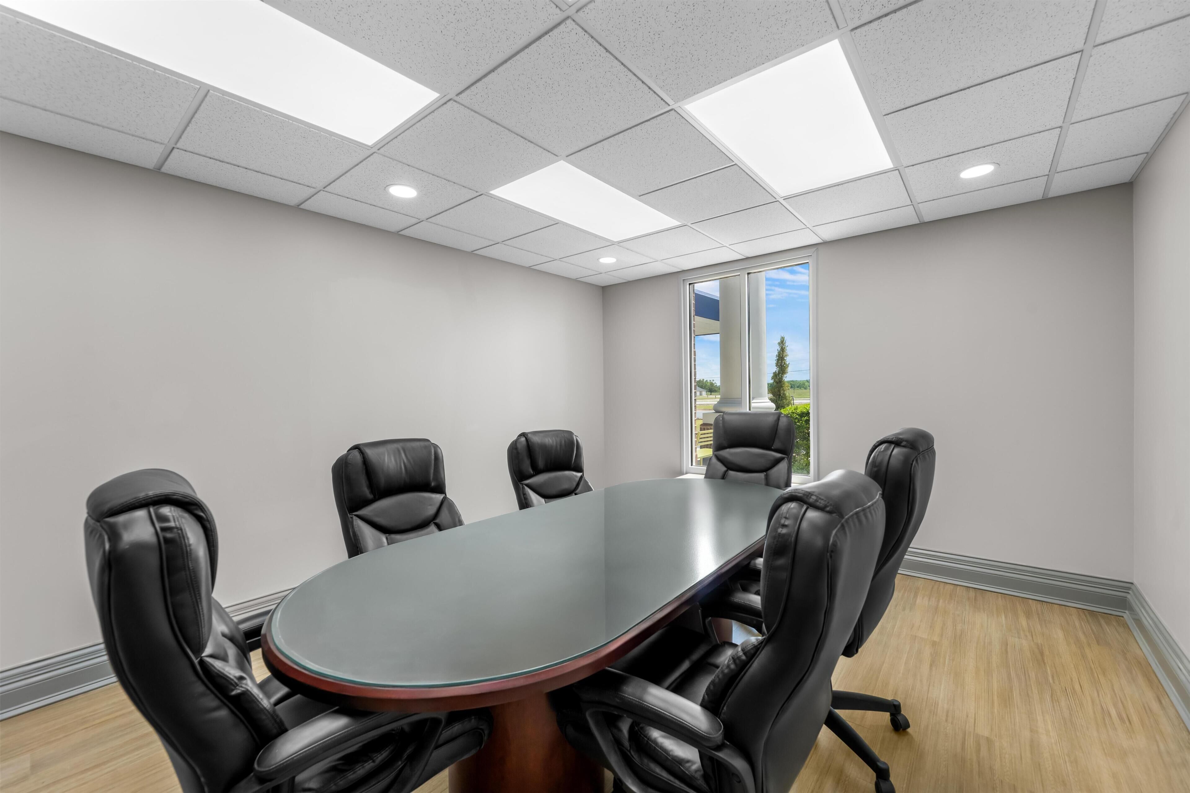 A conference room with a large oval table, black leather chairs, white walls, and a window with a view of trees.