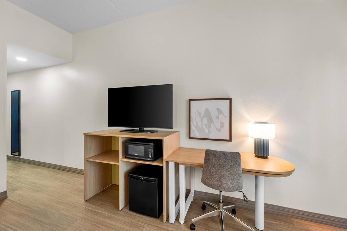 Habitación de hotel con escritorio de madera, televisor, microondas, cesto de basura, una silla y un cuadro en la pared.