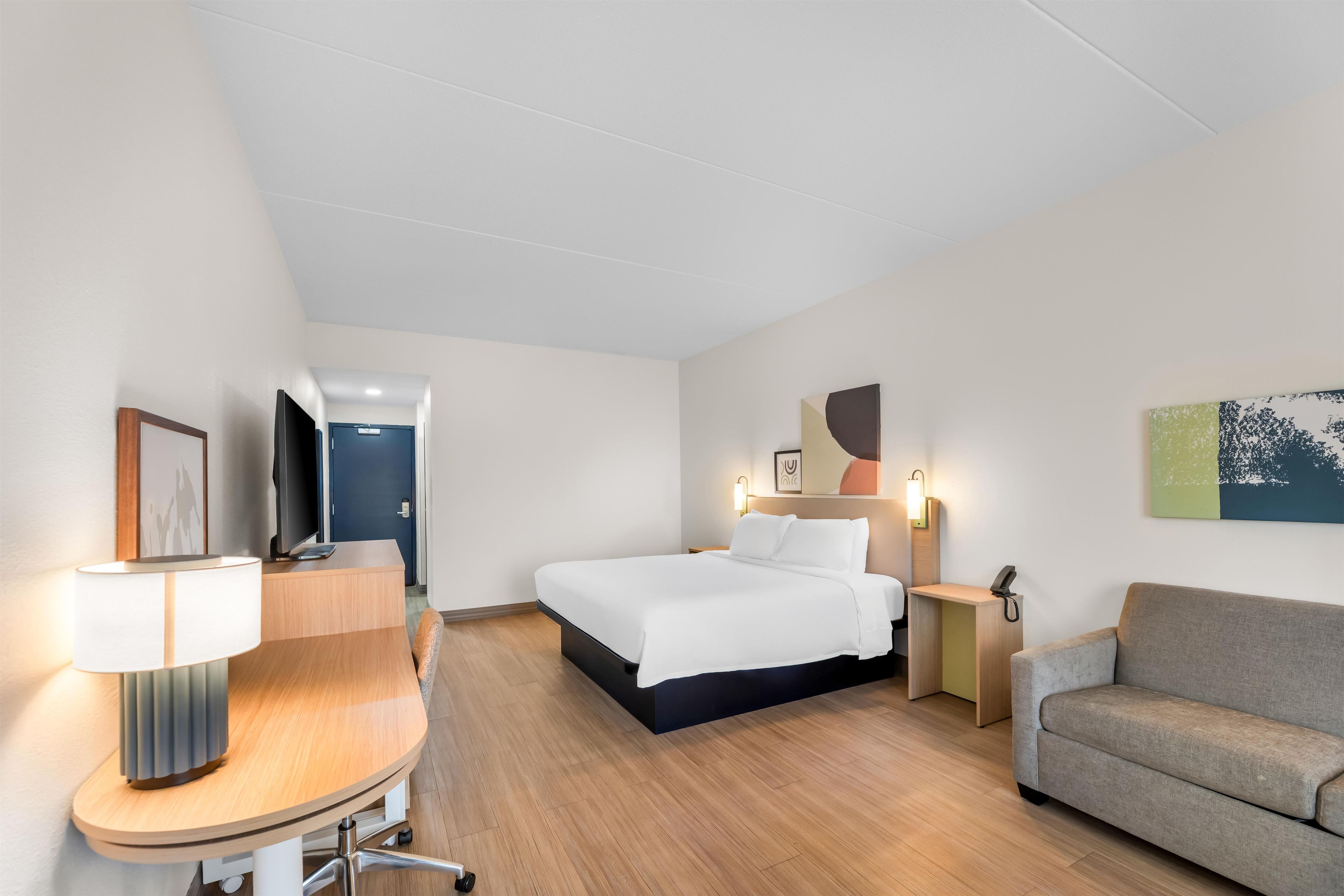 A hotel room with a bed, a desk, a chair, a sofa, a desk lamp, a bedside table, a phone, and a wall-mounted artwork.