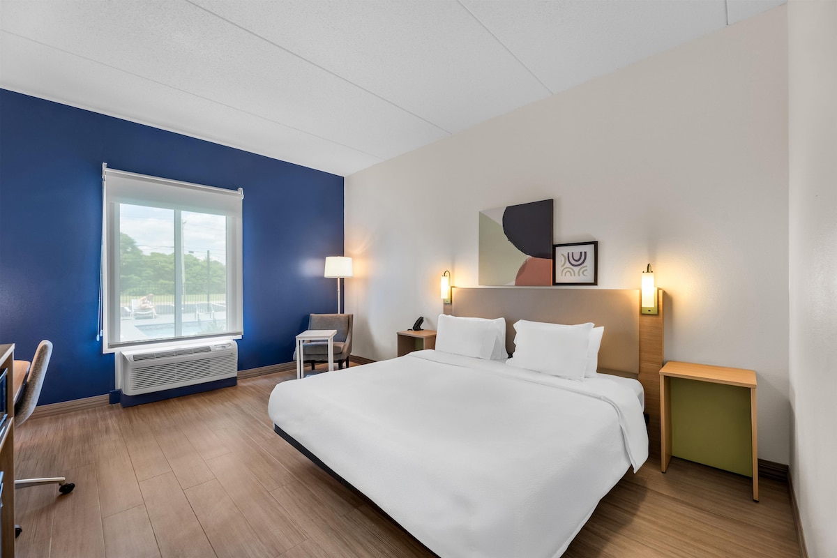 A hotel room with a large bed, a desk, a chair, a window, and a wall-mounted artwork.