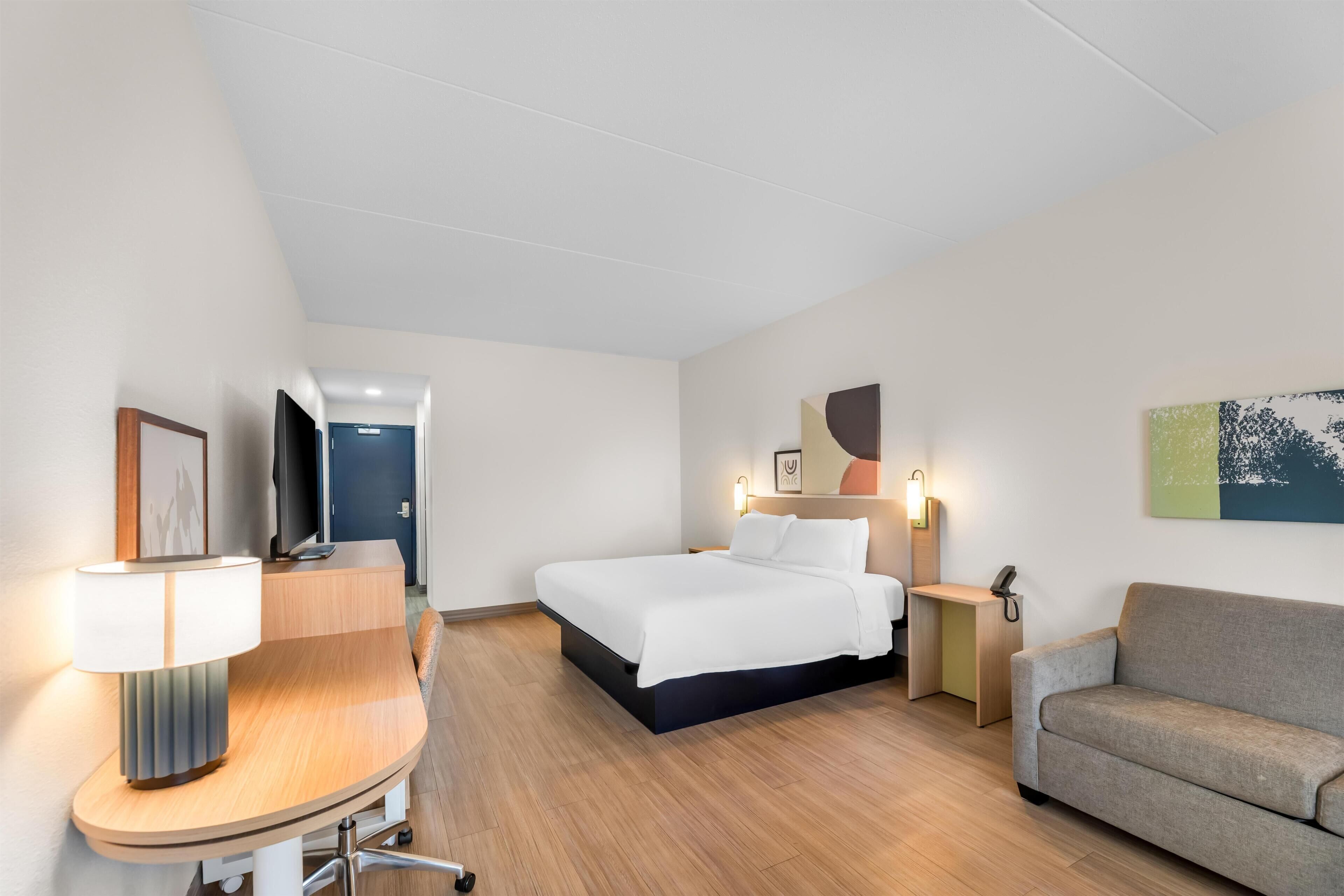 Habitación de hotel con cama, escritorio, silla, sofá, lámpara de escritorio, mesita de noche, teléfono y una obra de arte colgada en la pared.