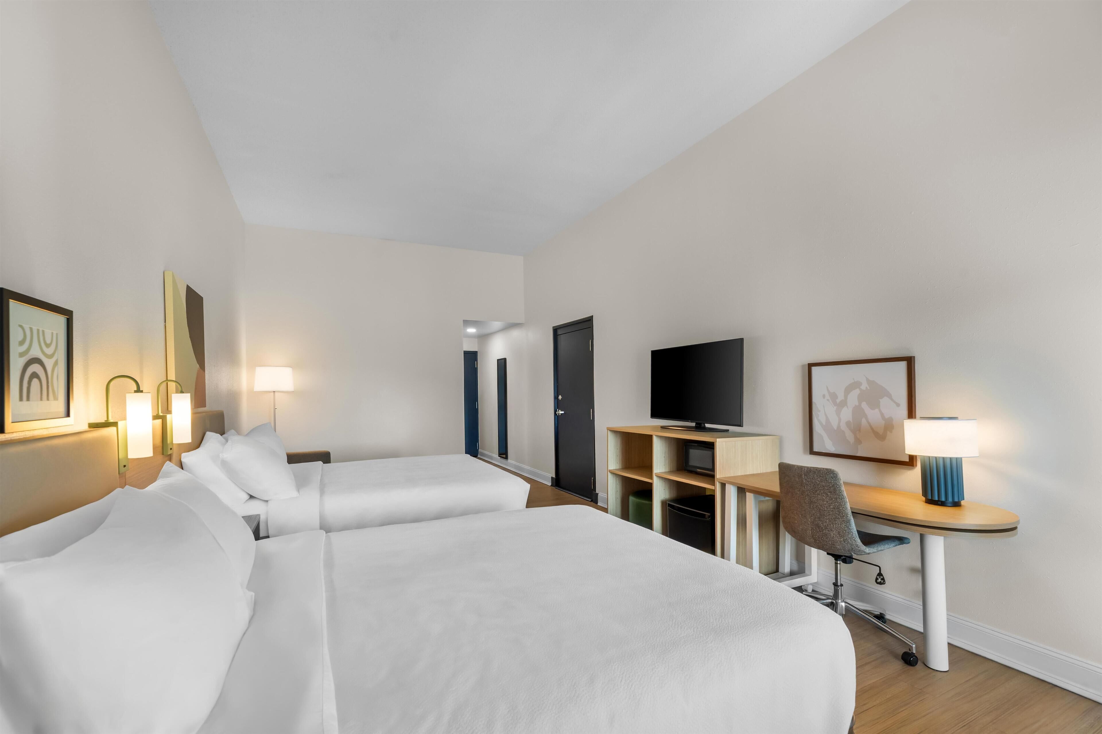 A hotel room with two beds, a desk with a chair, a television, and wall-mounted lamps.