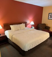 Habitación de hotel con una cama grande, una mesita de noche, una lámpara, una silla y un cuadro en la pared.
