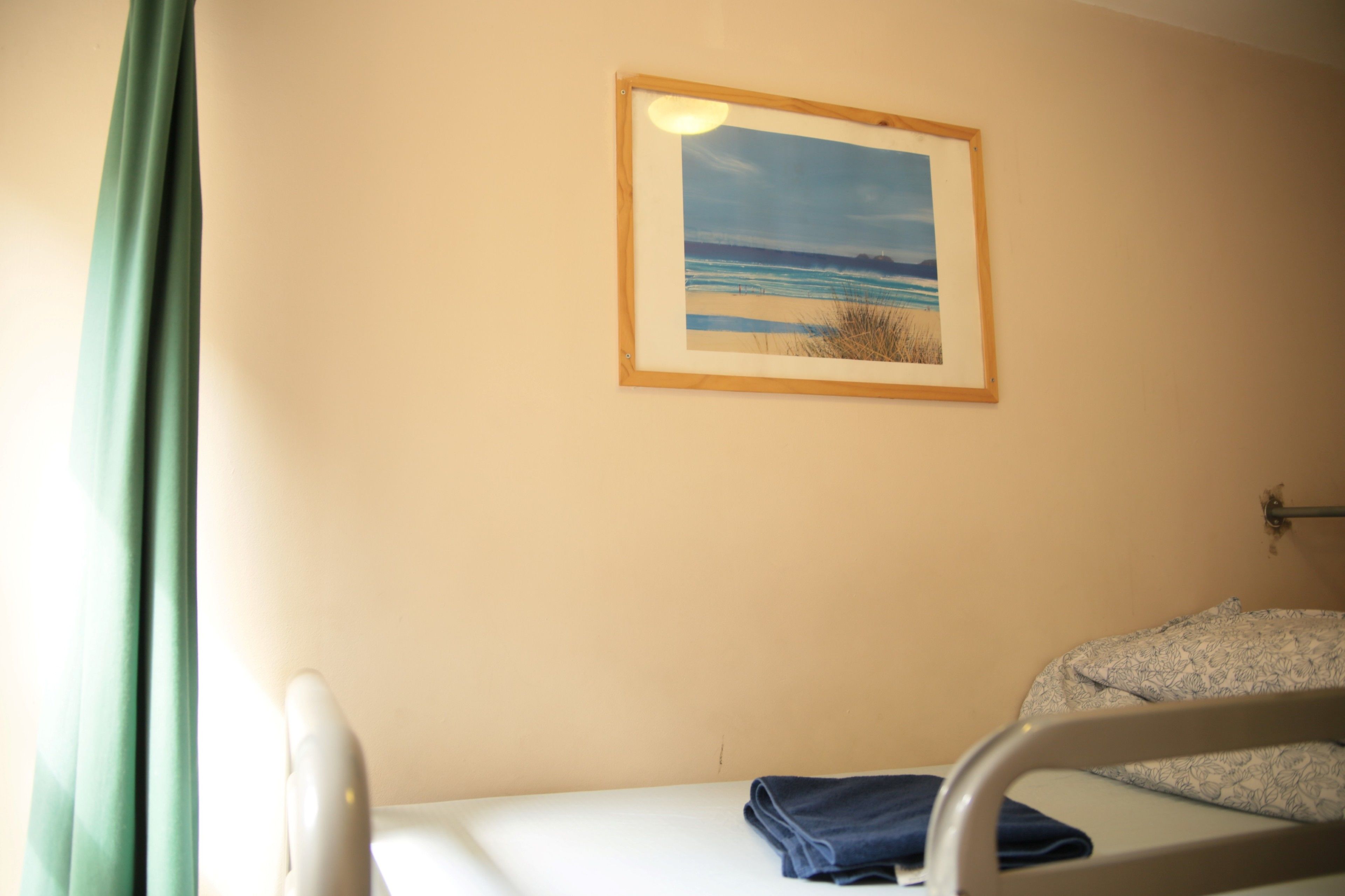 A single bed with a blue blanket, a framed picture of a beach scene, and a green curtain.