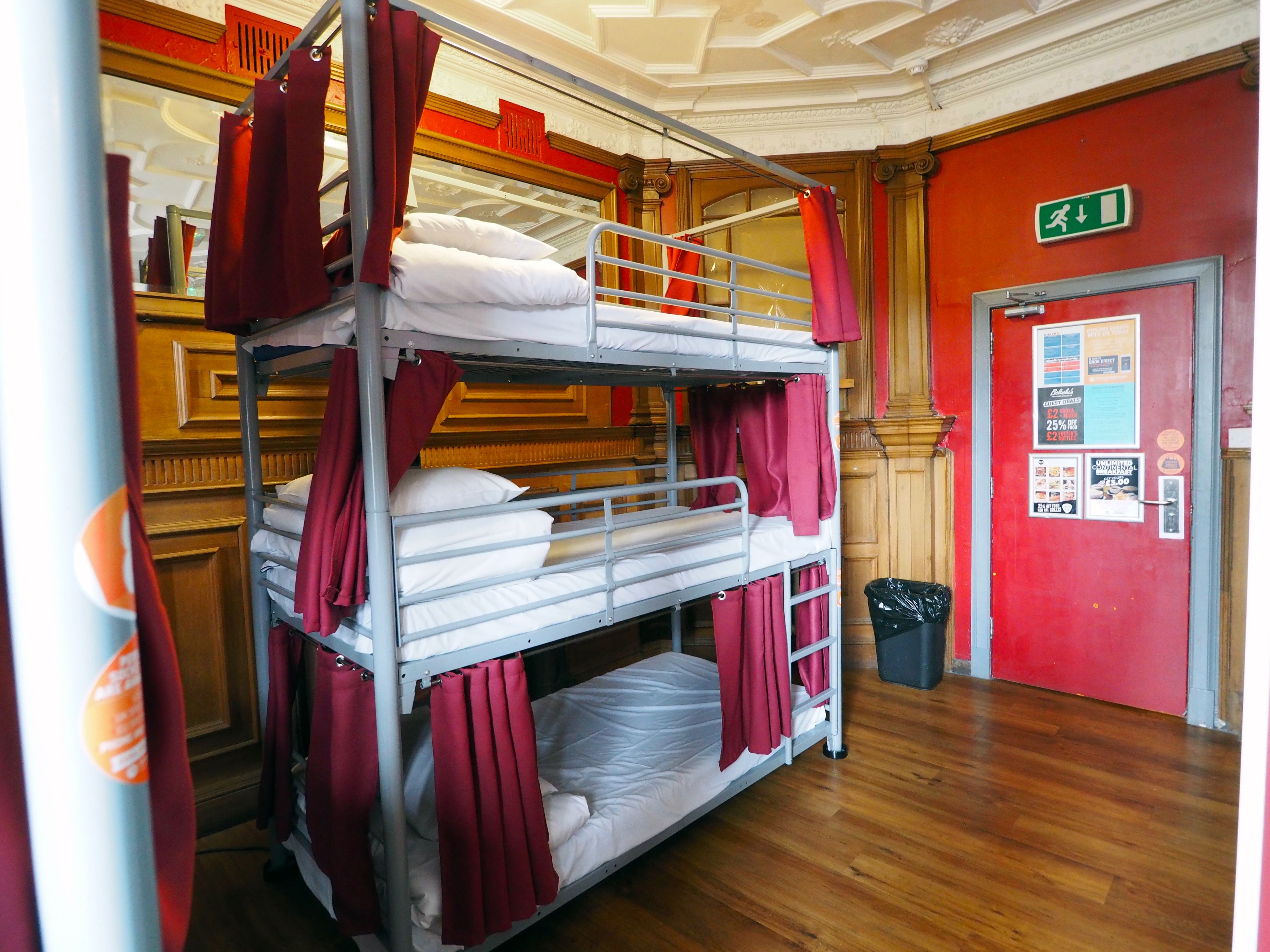 Une chambre avec des lits superposés, des rideaux rouges, un plancher en bois et une porte portant un panneau indiquant la sortie de secours.