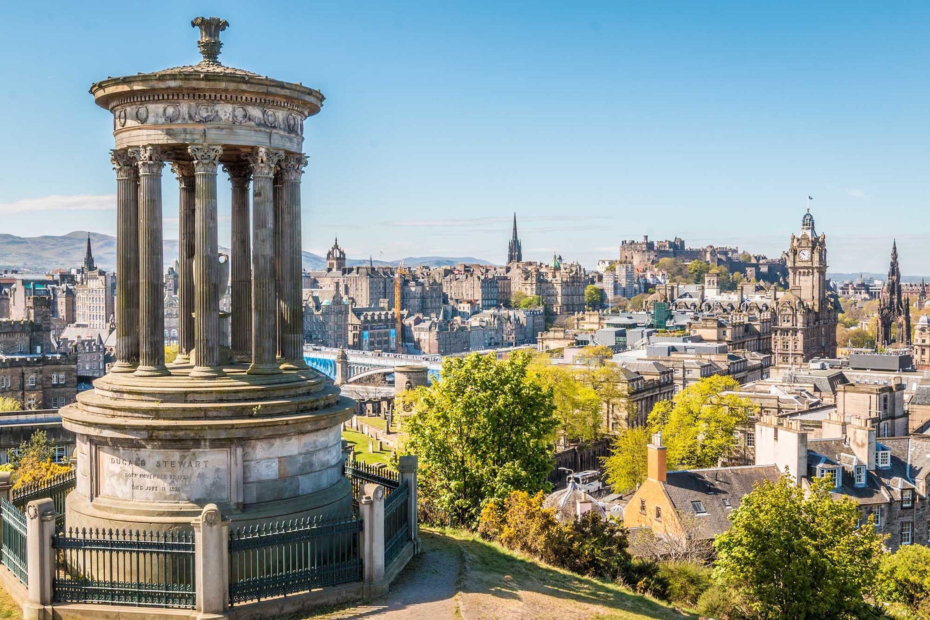 Ein historisches Denkmal mit Inschriften, umgeben von einem grünen Zaun, vor einer Stadtlandschaft mit verschiedenen Gebäuden und blauem Himmel.