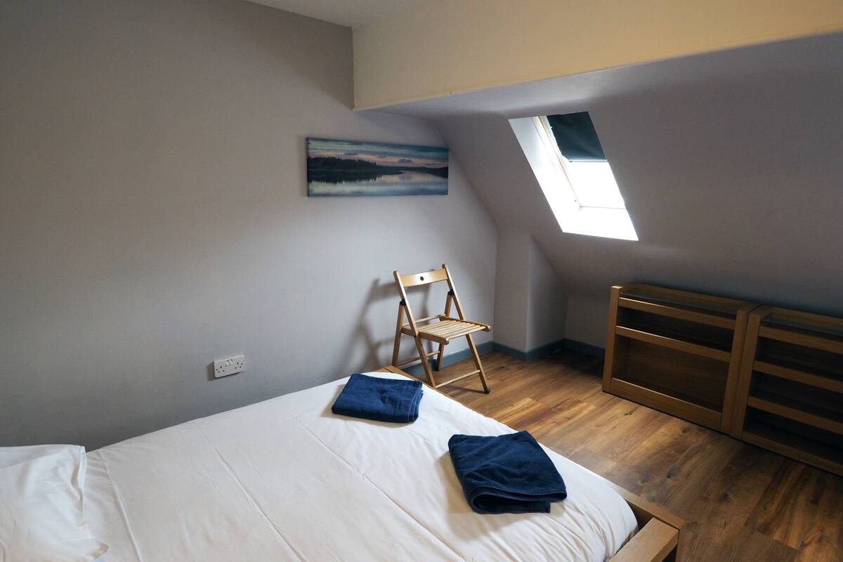 A bedroom with a bed, a wooden chair, a bench, and a skylight.