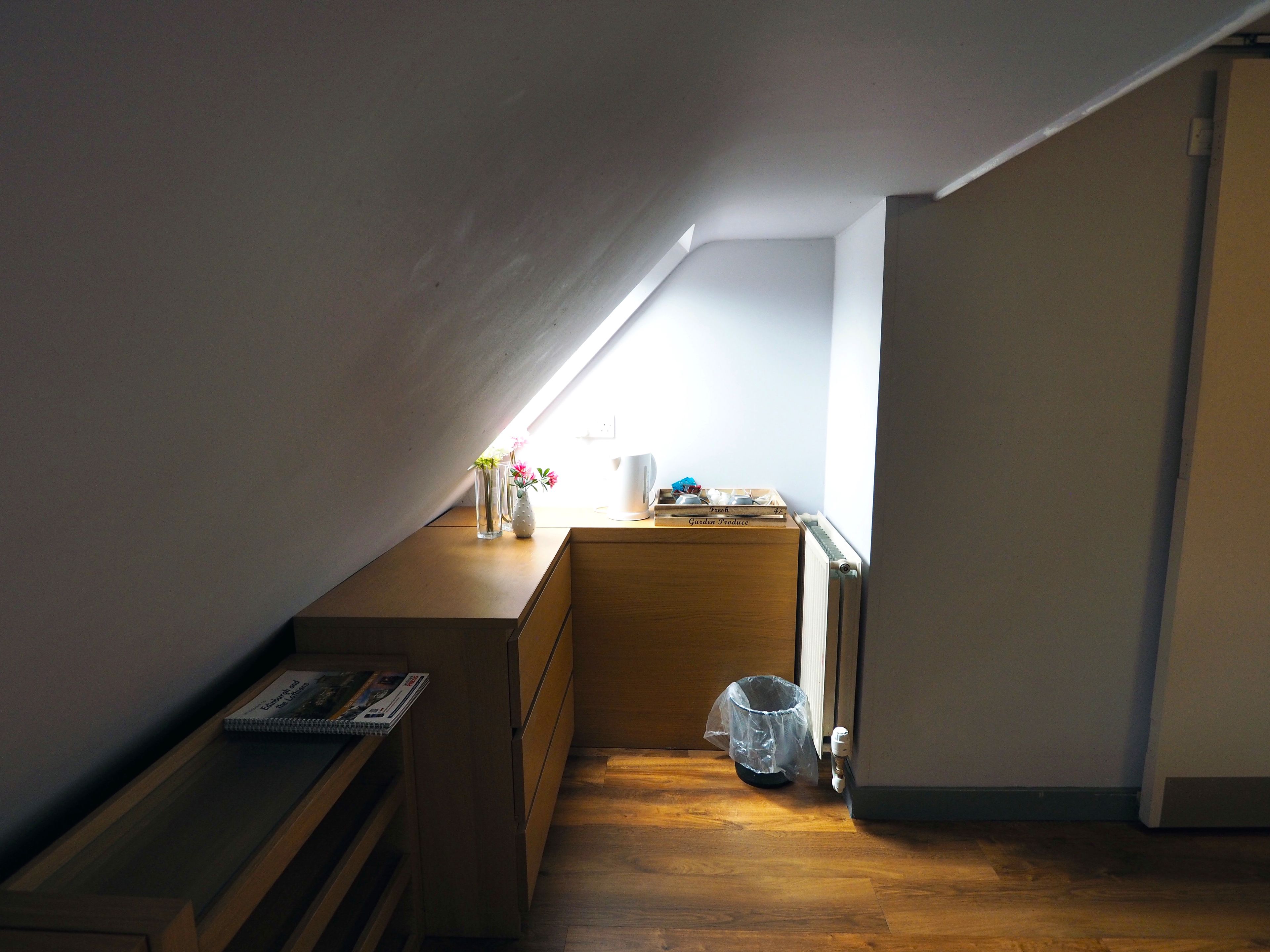 Quarto sótão pequeno com teto inclinado, soalho de madeira, secretária de madeira, radiador e janela que proporciona luz natural.