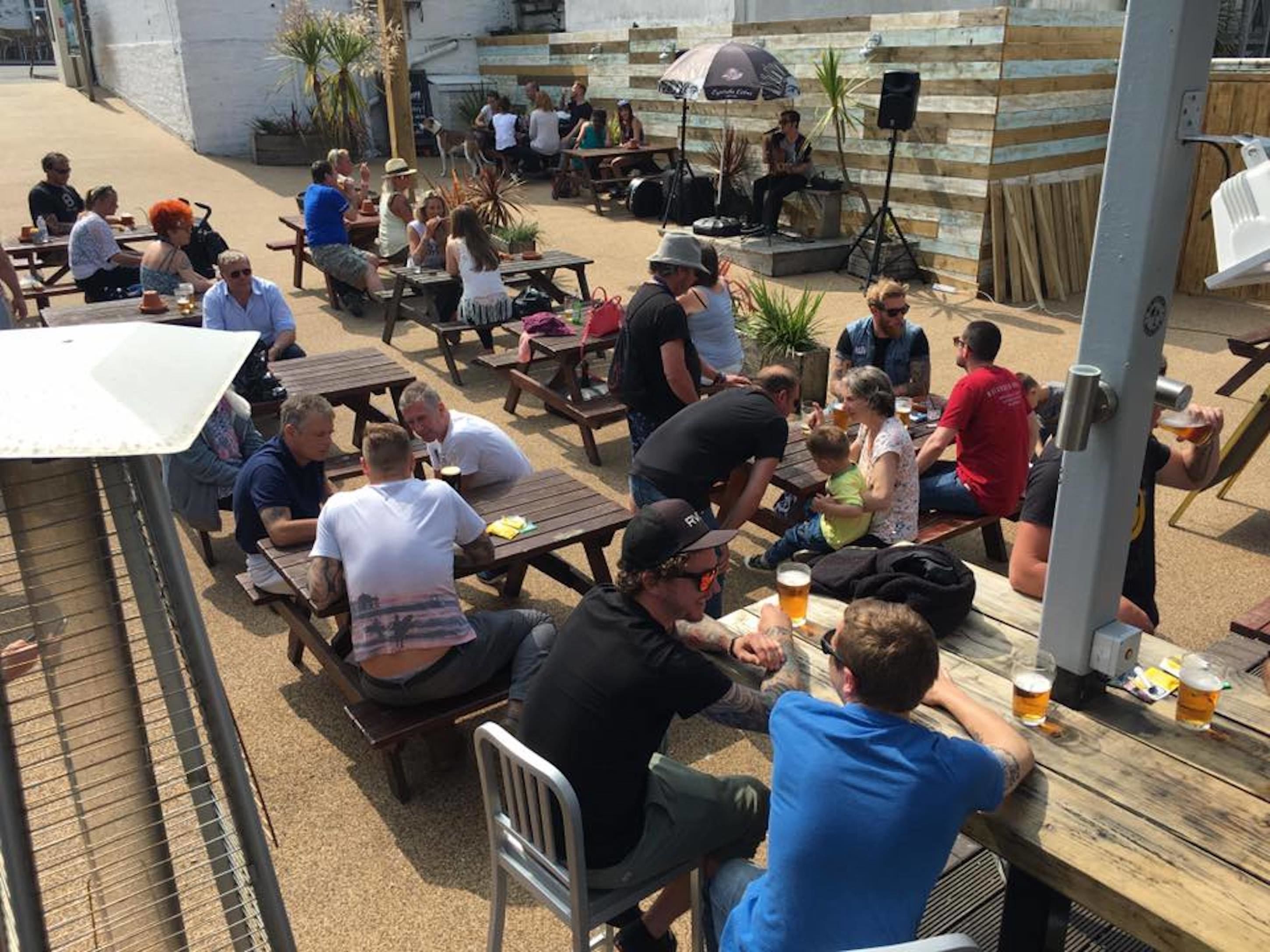 Een terras buiten met houten tafels en banken, waarvan sommigen bekleed zijn met zittende mensen en anderen staan.