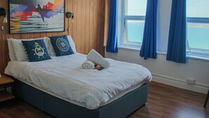 A neatly made bed with a white comforter and blue pillows, a wooden headboard, a painting of a boat on water, and a window with blue curtains. - St Christopher's Inn, Newquay - Hostel (Newquay)