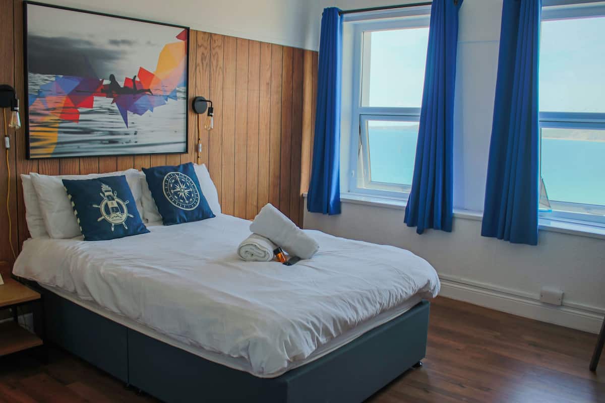 A neatly made bed with a white comforter and blue pillows, a wooden headboard, a painting of a boat on water, and a window with blue curtains.