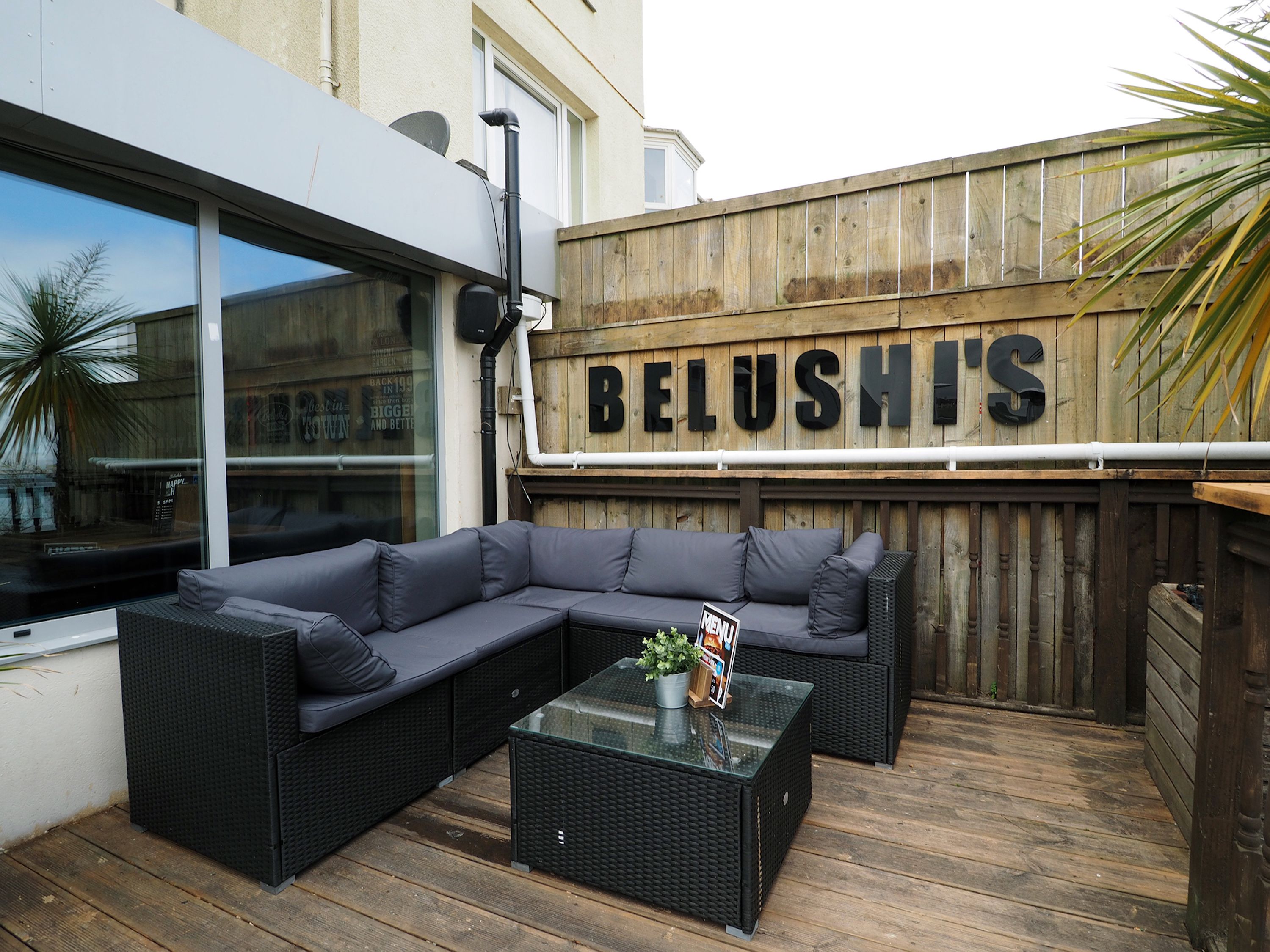 An outdoor seating area with a large sectional sofa and a glass-top coffee table, a wooden fence with the name "BELUSH'S" on it, and a building with large windows in the background.