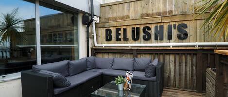 An outdoor seating area with a large sectional sofa and a glass-top coffee table, a wooden fence with the name "BELUSH'S" on it, and a building with large windows in the background.