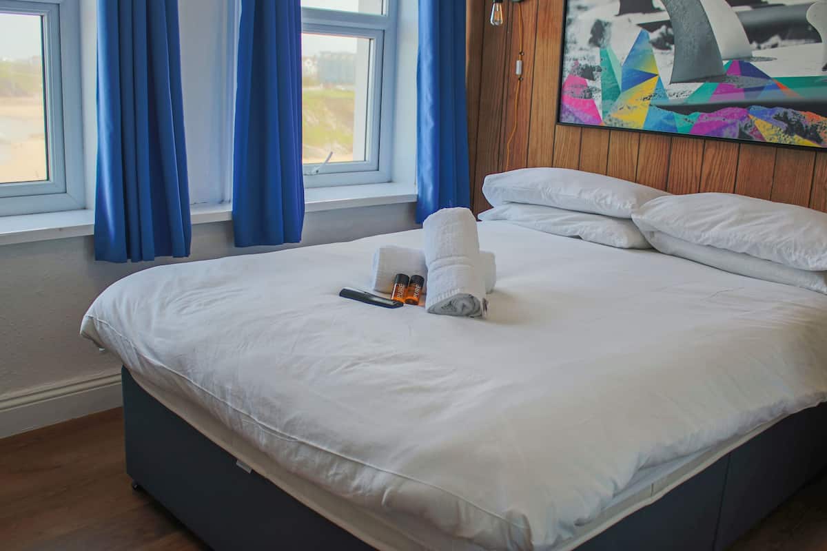 A neatly made bed with white linens, a remote control, and a rolled towel on a dark-colored bed frame.