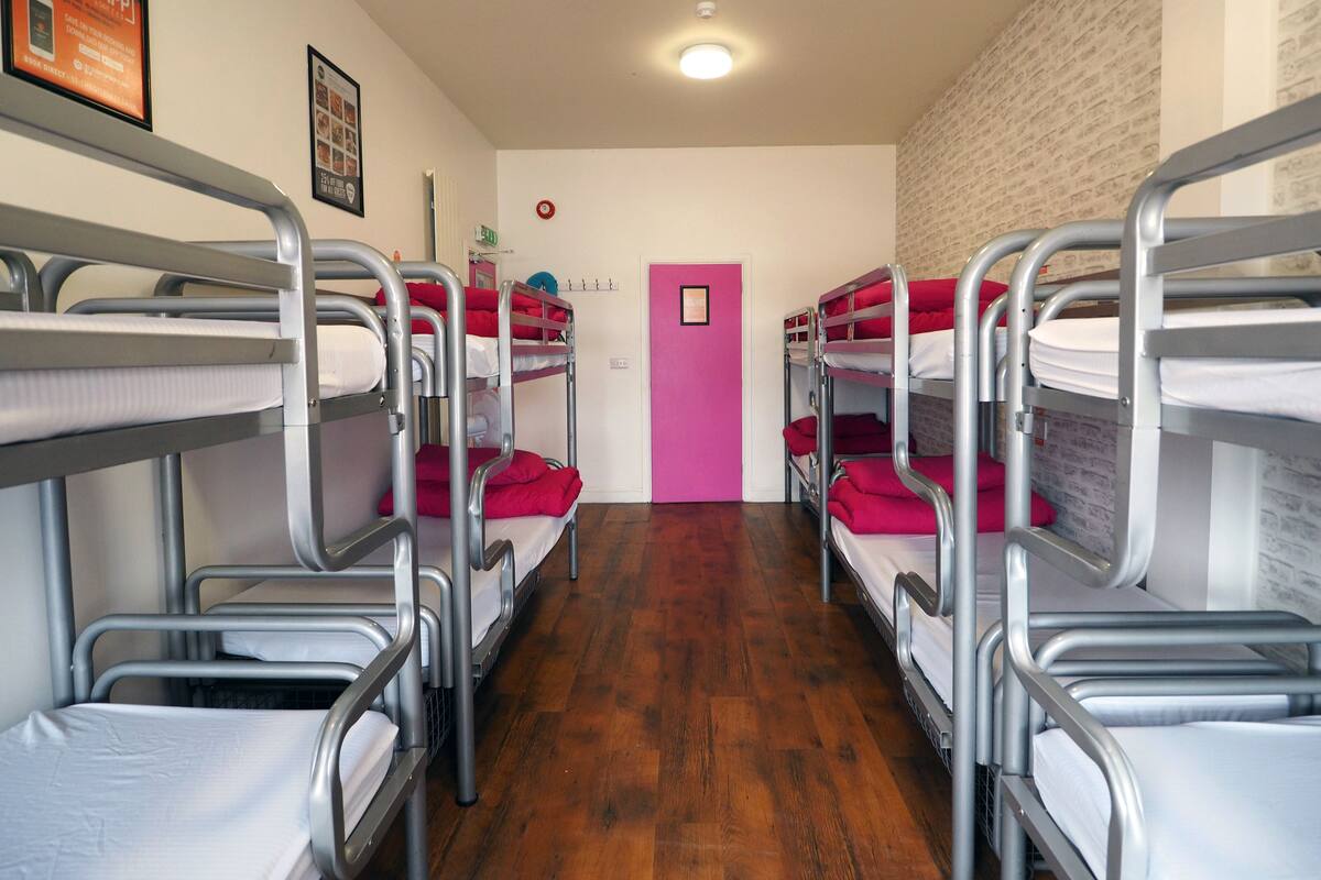 A dormitory room with bunk beds, wooden flooring, a pink door, and a framed picture on the wall.